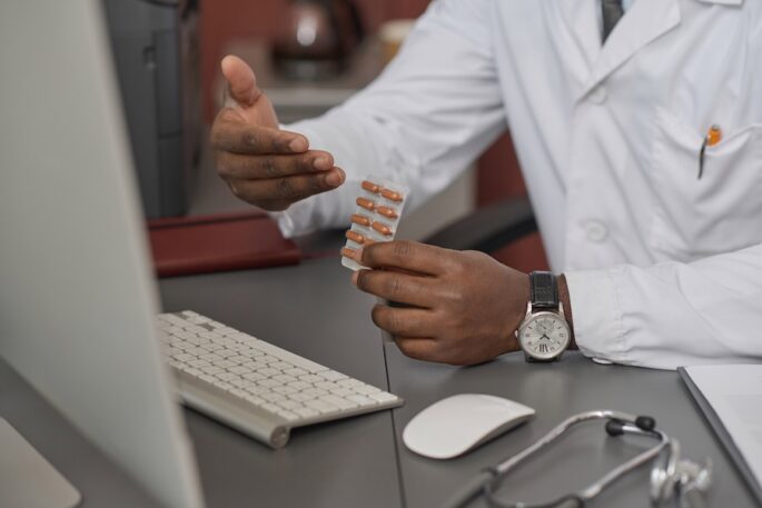 Hands of doctor showing medicine in capsules to computer webcam during video call with patient