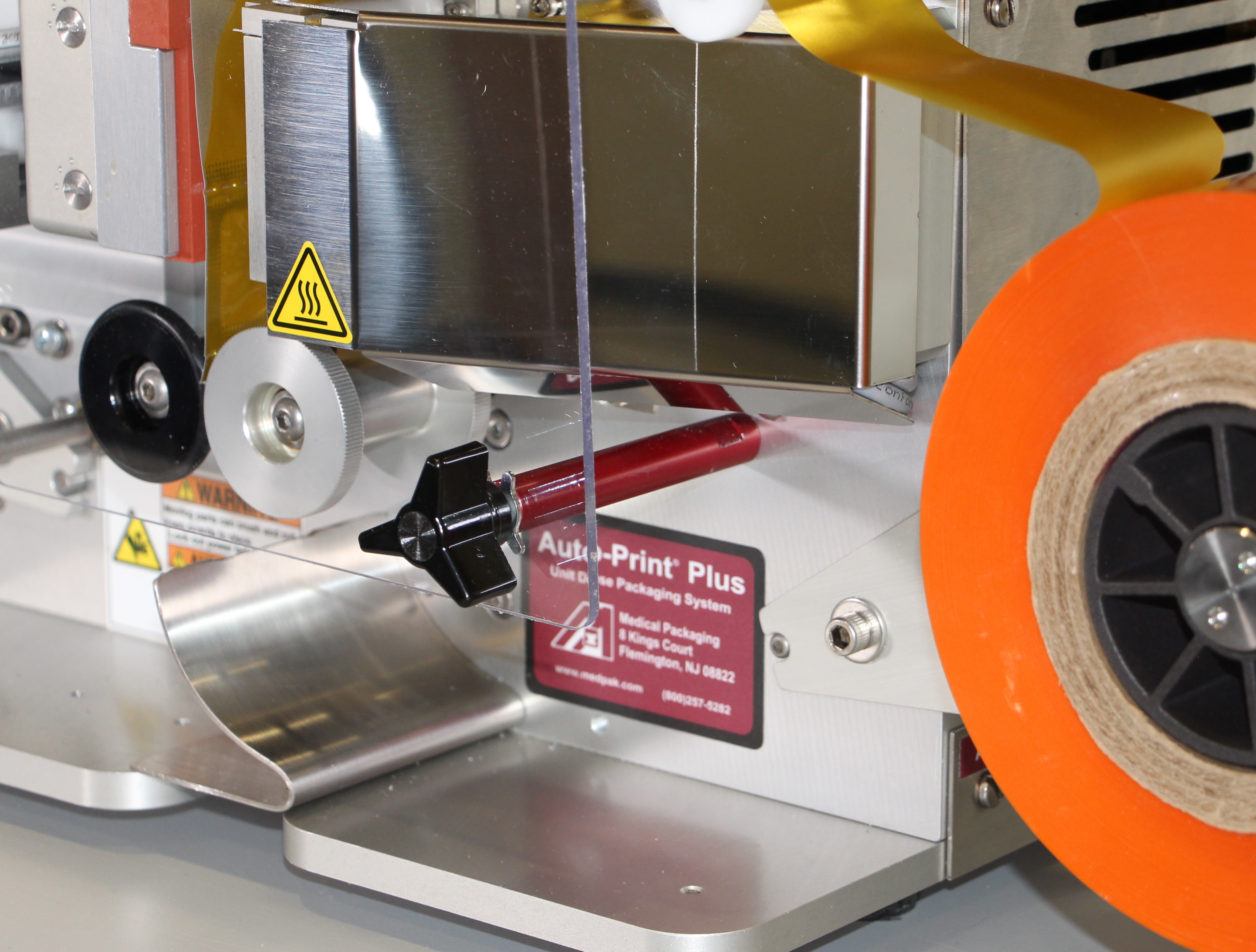 Close-up of an industrial labeling machine with an orange label roll and metal rollers used to apply labels to medical packaging.